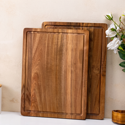 Three rectangular wooden cutting boards with rounded corners and raised edges, stacked slightly offset against a beige wall. A small vase with white flowers sits on the right side of the frame.