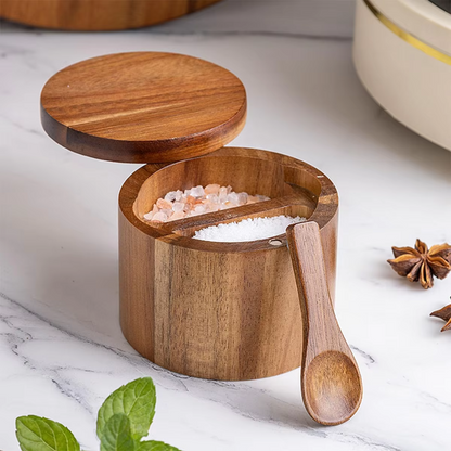 A wooden salt cellar with a spoon, containing white salt, placed on a kitchen countertop with a marble background. The wood has a natural brown finish and the spoon has a light wood handle.