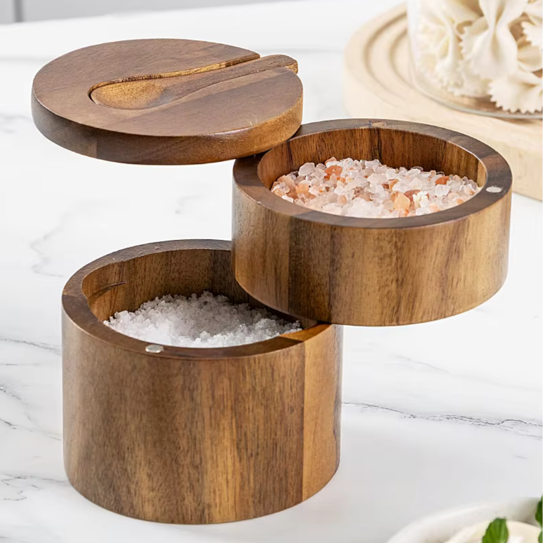 A wooden salt cellar with a spoon, containing white salt, placed on a kitchen countertop with a marble background. The wood has a natural brown finish and the spoon has a light wood handle.