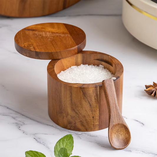 A wooden salt cellar with a spoon, containing white salt, placed on a kitchen countertop with a marble background. The wood has a natural brown finish and the spoon has a light wood handle.