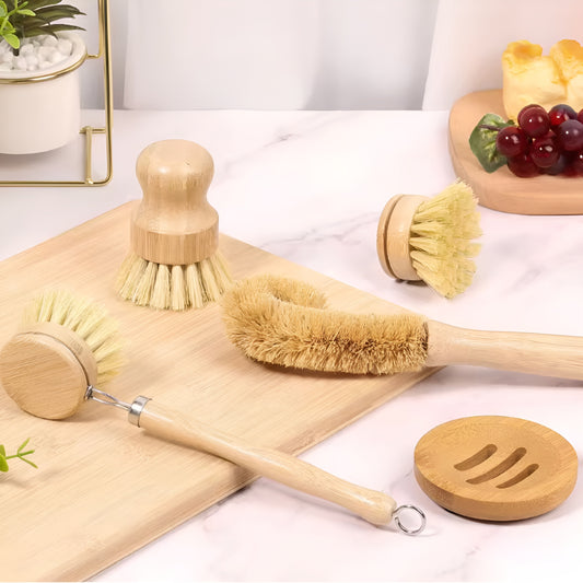 Set of wooden kitchen cleaning brushes and a soap dish on a light surface with a plant and fruit in the background.
