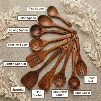 A set of wooden kitchen utensils arranged in a fan shape on a textured beige fabric background with decorative dried leaves. The utensils include labeled pieces: rice spoon, salad spoon, mixing spoon, strainer spoon, slotted spatula, spatula, flat spatula, spaghetti spoon, salad fork, and soup ladle.