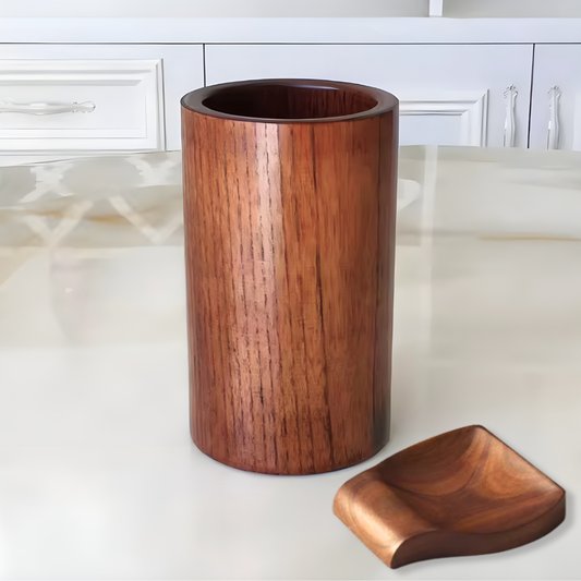 A cylindrical wooden cup or holder with a rich reddish-brown wood grain finish, shown alongside a small matching wooden tray on a light-colored countertop with white kitchen cabinets in the background.